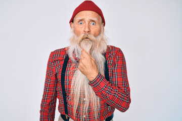 Old senior man with grey hair and long beard wearing hipster look with wool cap looking fascinated with disbelief, surprise and amazed expression with hands on chin