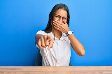 Beautiful hispanic woman wearing casual clothes sitting on the table laughing at you, pointing finger to the camera with hand over mouth, shame expression