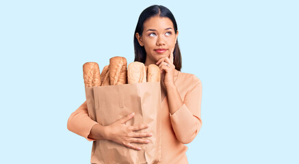 Young beautiful latin girl holding paper bag with bread serious face thinking about question with...