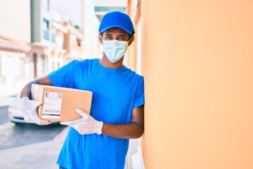 Obraz na płótnie Canvas African delivery man wearing courier uniform outdoors wearing coronavirus safety mask holding cardboard parcel