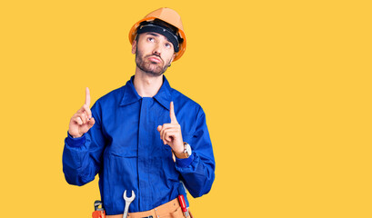 Young hispanic man wearing worker uniform pointing up looking sad and upset, indicating direction with fingers, unhappy and depressed.
