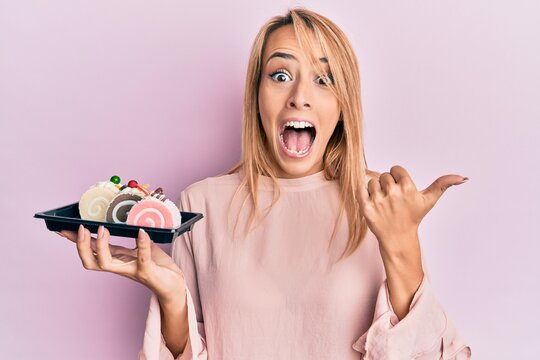 Beautiful blonde woman holding cake sweets pointing thumb up to the side smiling happy with open mouth