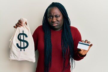 Young black woman with braids holding dollars bag and credit card clueless and confused expression....
