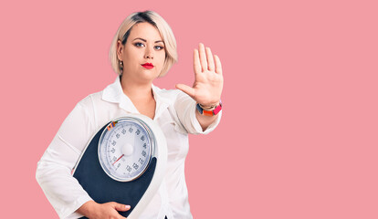 Young blonde plus size woman holding weighing machine with open hand doing stop sign with serious...