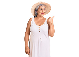 Senior woman with gray hair wearing summer hat and summer clothes smiling with happy face looking and pointing to the side with thumb up.
