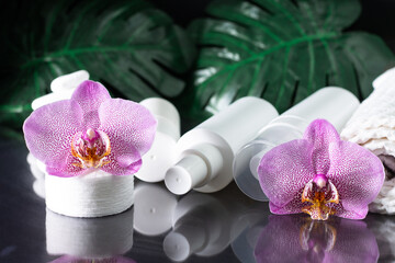 Lilac orchid flower, white cosmetic bottles, cotton pads with stack of white stones and monstera leaves on black surface