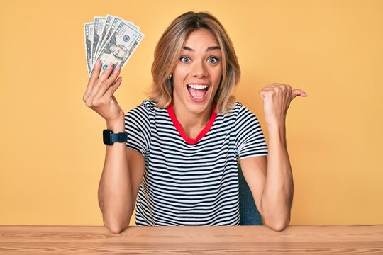 Beautiful caucasian woman holding 20 dollars banknotes pointing thumb up to the side smiling happy with open mouth