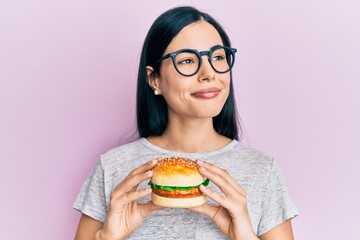 Beautiful young woman eating tasty hamburger smiling looking to the side and staring away thinking.