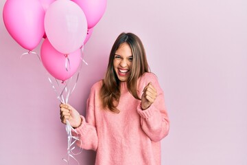 Young brunette woman holding pink balloons screaming proud, celebrating victory and success very...