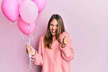 Fotobehang Young brunette woman holding pink balloons screaming proud, celebrating victory and success very excited with raised arms © Krakenimages.com