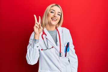 Young caucasian woman wearing doctor uniform and stethoscope showing and pointing up with fingers number two while smiling confident and happy.