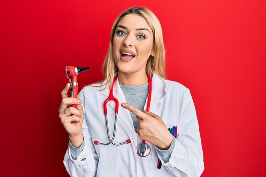 Young caucasian woman wearing doctor uniform holding otoscope smiling happy pointing with hand and finger