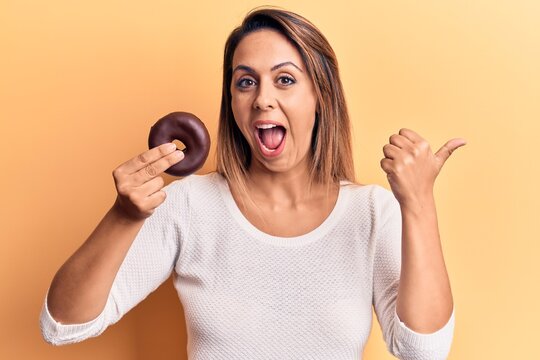 Young beautiful woman holding donut pointing thumb up to the side smiling happy with open mouth