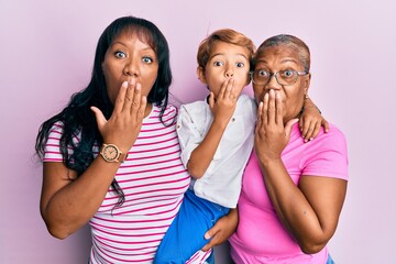 Hispanic family of grandmother, mother and son hugging together covering mouth with hand, shocked...