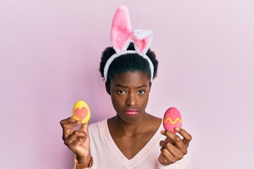 Young african american girl wearing cute easter bunny ears holding painted eggs depressed and worry...