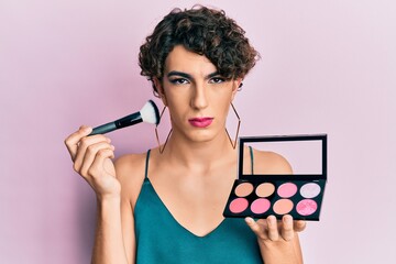Young man wearing woman make up holding makeup brush and blush skeptic and nervous, frowning upset...
