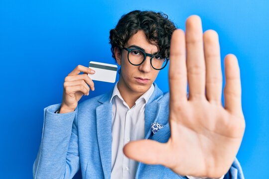 Hispanic young man holding credit card with open hand doing stop sign with serious and confident expression, defense gesture