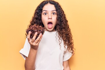 Beautiful kid girl with curly hair holding chocolate cereals scared and amazed with open mouth for surprise, disbelief face