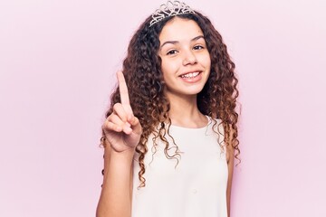 Beautiful kid girl with curly hair wearing princess tiara smiling with an idea or question pointing finger up with happy face, number one © Krakenimages.com