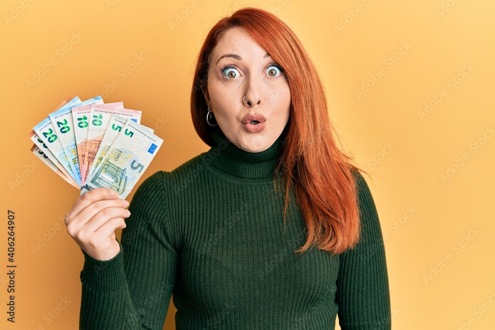 Poster beautiful redhead woman holding euro banknotes scared and amazed with open mouth for surprise, disbe