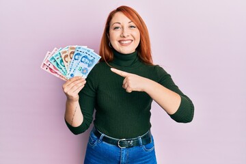 Beautiful redhead woman holding yuan chinese banknotes smiling happy pointing with hand and finger