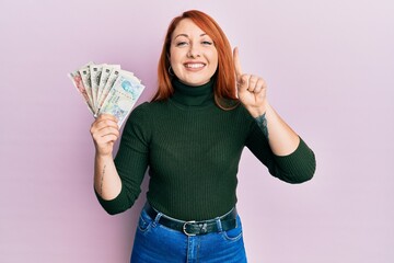 Beautiful redhead woman holding united kingdom pounds smiling with an idea or question pointing finger with happy face, number one