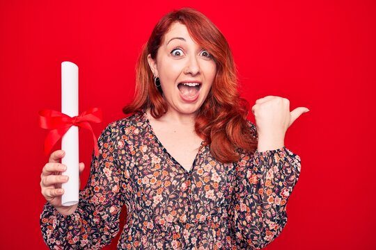 Young beautiful redhead woman holding graduated degree diploma over red background pointing thumb up to the side smiling happy with open mouth