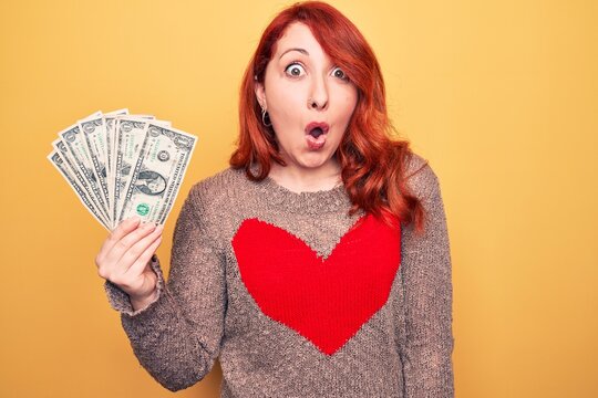Young beautiful redhead woman holding bunch of dollars banknotes over yellow background scared and amazed with open mouth for surprise, disbelief face