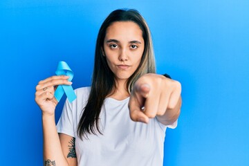Young hispanic woman holding blue ribbon pointing with finger to the camera and to you, confident...