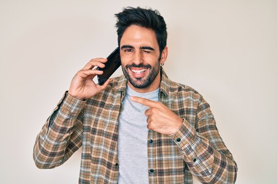 Handsome hispanic man with beard having conversation talking on the smartphone winking looking at the camera with sexy expression, cheerful and happy face.