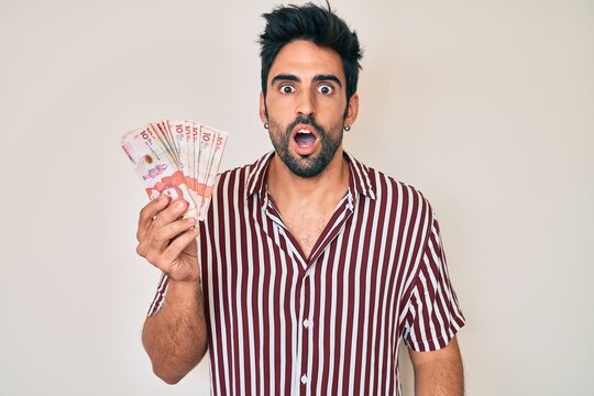 Handsome hispanic man with beard holding colombian pesos scared and amazed with open mouth for surprise, disbelief face