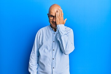 Middle age bald man wearing casual clothes and glasses covering one eye with hand, confident smile on face and surprise emotion.