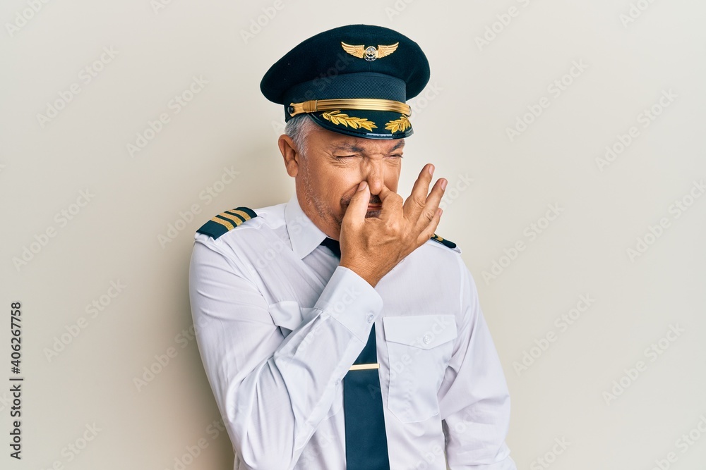 Poster Handsome middle age mature man wearing airplane pilot uniform smelling something stinky and disgusting, intolerable smell, holding breath with fingers on nose. bad smell