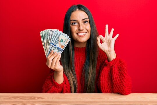 Beautiful hispanic woman holding dollars doing ok sign with fingers, smiling friendly gesturing excellent symbol