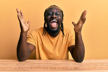 Handsome young black man wearing casual clothes sitting on the table celebrating mad and crazy for...