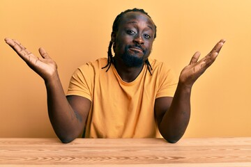 Handsome young black man wearing casual clothes sitting on the table clueless and confused...