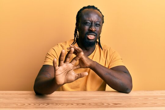 Handsome young black man wearing casual clothes sitting on the table disgusted expression, displeased and fearful doing disgust face because aversion reaction. with hands raised