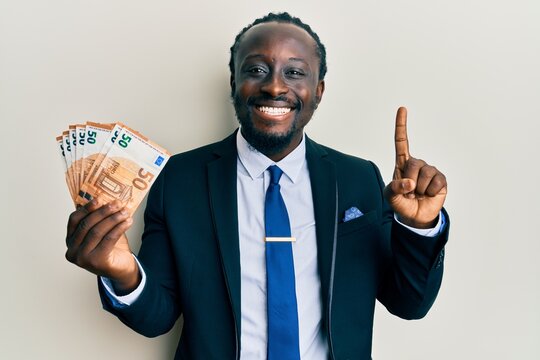 Handsome young black man wearing business suit holding 50 euros banknotes smiling with an idea or question pointing finger with happy face, number one