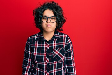 Young hispanic woman with curly hair wearing casual clothes and glasses puffing cheeks with funny...