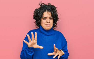 Young hispanic woman with curly hair wearing turtleneck sweater disgusted expression, displeased and fearful doing disgust face because aversion reaction.