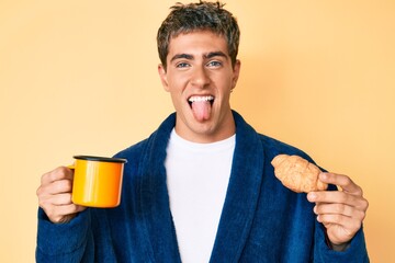 Young handsome man wearing bathrobe eating breakfast holding coffee and croissant sticking tongue out happy with funny expression.