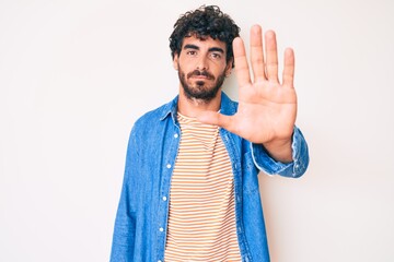 Handsome young man with curly hair and bear wearing casual denim jacket doing stop sing with palm of the hand. warning expression with negative and serious gesture on the face.