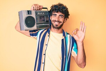 Handsome young man with curly hair and bear holding boombox, listening to music wearing summer look doing ok sign with fingers, smiling friendly gesturing excellent symbol