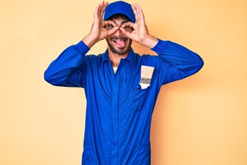 Handsome young man with curly hair and bear wearing builder jumpsuit uniform doing ok gesture like binoculars sticking tongue out, eyes looking through fingers. crazy expression.