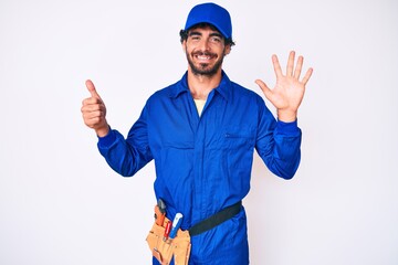 Handsome young man with curly hair and bear weaing handyman uniform showing and pointing up with fingers number six while smiling confident and happy.