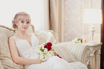 Pretty young Bride in elegant wedding dress with her bouquet. Blonde-haired woman with wedding hair-style in royal room of hotel. Boudoir morning of the bride in luxury interior. Wedding day ceremony