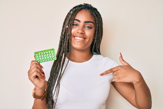 Young african american woman with braids holding birth control pills pointing finger to one self smiling happy and proud