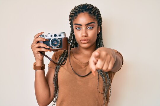 Young african american woman with braids holding vintage camera pointing with finger to the camera and to you, confident gesture looking serious