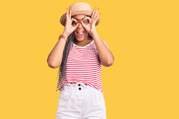 Young african american woman with braids wearing summer hat doing ok gesture like binoculars sticking tongue out, eyes looking through fingers. crazy expression.