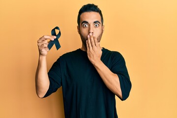 Young hispanic man holding black ribbon covering mouth with hand, shocked and afraid for mistake....
